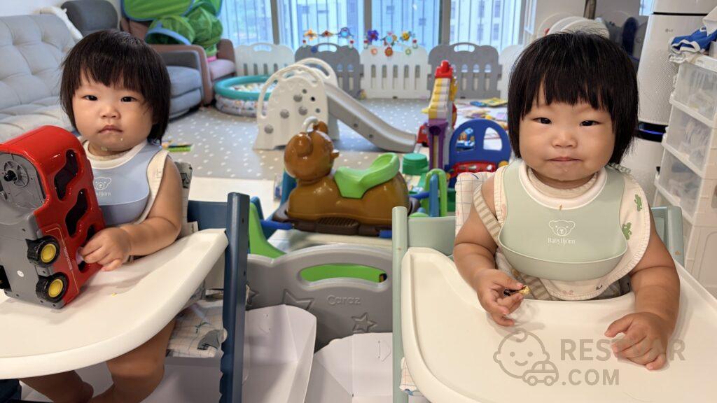 하이체어 두 대에 나란히 앉아 각자 베이비뵨 실리콘 턱받이를 한 쌍둥이