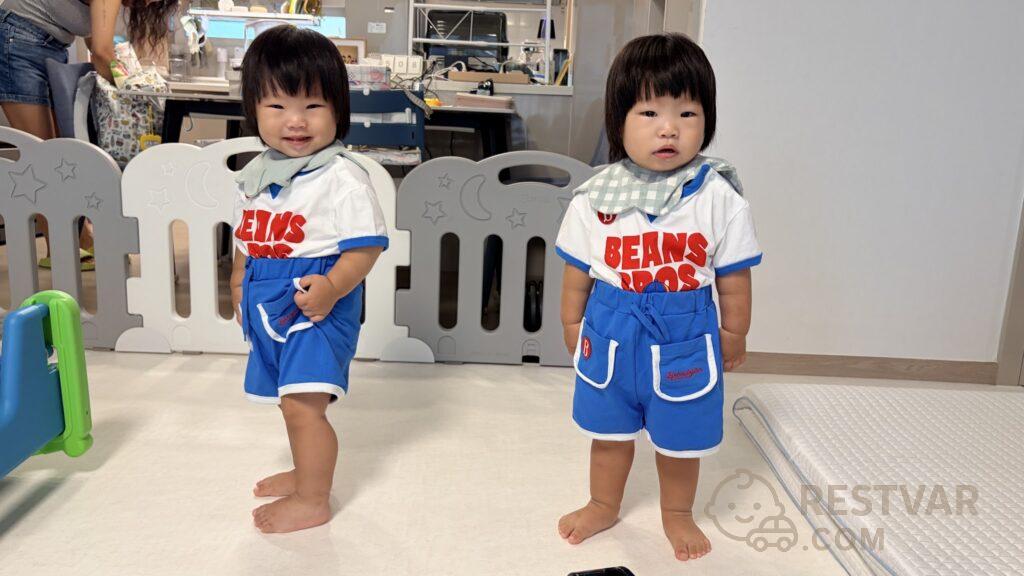 외출복을 입고 집 거실에 서 있는 16개월 쌍둥이